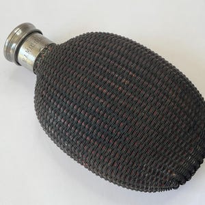 Puede incluir: Un frasco antiguo con un exterior tejido de color marrón oscuro y una tapa de metal plateado. El frasco tiene una forma oblonga redondeada. La tapa está grabada con el texto "W.A. MACLELLAN". El frasco es probablemente de principios del siglo XX.