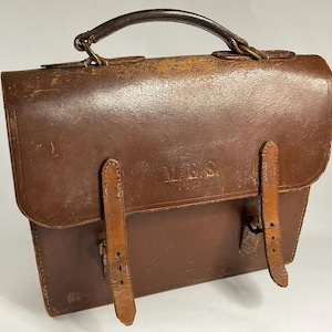 以下が含まれることがあります： トップハンドルと2つのフロントストラップが付いた、ヴィンテージの茶色の革製サッチェル。サッチェルは、革に目に見える摩耗や損傷があり、使い古された外観をしています。「M.E.S.」のイニシャルが前面にエンボス加工されています。