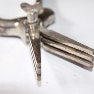 Op de afbeelding: Een zilveren metalen gereedschap met een handvat en meerdere bladen. Het gereedschap heeft een kleine, ronde knop op het handvat.