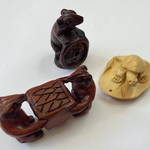 Puede incluir: Tres figuras de madera tallada: un oso con una moneda, dos osos que llevan una caja y una tortuga sobre una roca. Las figuras de oso son de color marrón oscuro, mientras que la tortuga y la roca son de color marrón claro. La caja tiene un diseño texturizado detallado.