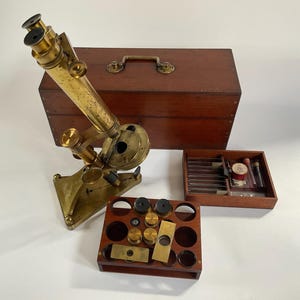May include: A vintage brass microscope with a triangular base, accompanied by a large wooden storage case with a metal handle, a wooden accessory tray containing glass slides and vials, and a wooden rack holding several brass lens components.