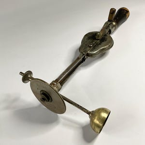 Puede incluir: Una cuchara para helado antigua con mango de madera y cuerpo metálico. La cuchara tiene un mecanismo de palanca y un disco circular. Los componentes metálicos de la cuchara muestran signos de edad y uso. El diseño general de la cuchara es de estilo vintage.