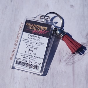 May include: A clear plastic keychain with a red tassel. The keychain features a black and white image of a Guardians of the Galaxy: Mission Breakout! FastPass reminder. The text on the keychain reads "FastPass Reminder. Please have your valid park admission available upon your return. Enter Between 7:50 PM and 8:50 PM. Another FastPass selection will be available after 7:41pm. Thu Jun 29 2017."