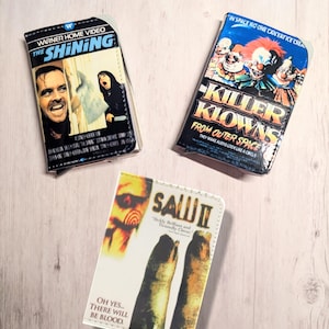 May include: Three wallets with movie poster designs. One wallet features "The Shining" with Jack Nicholson. Another displays "Killer Klowns From Outer Space." The third wallet shows "Saw II" with the text "Oh yes... There will be blood."