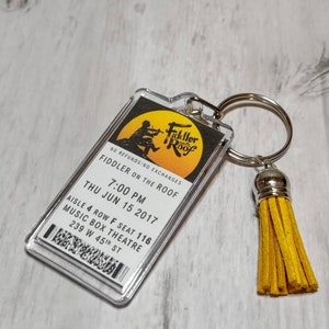 May include: A clear plastic keychain with a ticket stub for a performance of "Fiddler on the Roof" at the Music Box Theatre in New York City. The ticket stub shows the date as Thursday, June 15, 2017, at 7:00 PM. The keychain also has a yellow tassel.