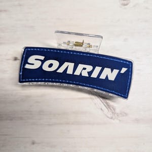 May include: A clear plastic hair claw clip with a navy blue rectangular patch. The patch features the word "SOARIN'" in white block letters. The clip has a metal spring mechanism.