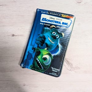 May include: A rectangular case featuring the Disney Pixar movie "Monsters, Inc." The case has a blue background with Sully and Mike Wazowski. Text includes "From the Creators of Toy Story," "Monsters, Inc.," and "An Instant Classic."