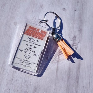 May include: A clear plastic keychain with a peach-colored tassel. The keychain features a printed ticket for "Universal's Action!" with the text "FASTPASS" and "Return Anytime Between 11:15 AM and 12:15 PM THU DEC 29 2011".