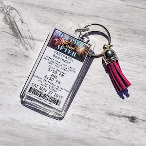 May include: A clear plastic keychain with a white card that says "Happily Ever After" with fireworks in the background. The card also says "Fastpass Reminder" and has instructions for park admission. The keychain has a pink tassel.