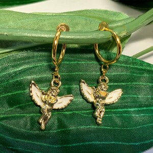 May include: A pair of gold hoop earrings with a white enamel angel charm hanging from each hoop. The angels are holding a heart in their hands.