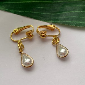 May include: A pair of gold-colored clip-on earrings. Each earring features a teardrop-shaped, faux pearl pendant encased in a gold-tone frame. The clip-on mechanism is also gold-colored.
