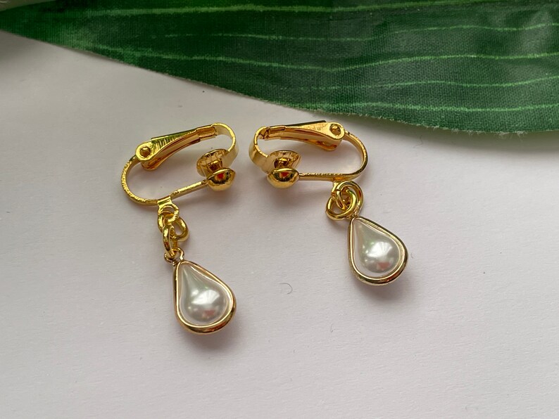 May include: A pair of gold-colored clip-on earrings. Each earring features a teardrop-shaped, faux pearl pendant encased in a gold-tone frame. The clip-on mechanism is also gold-colored.