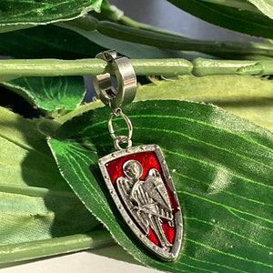 Op de afbeelding: Zilveren engelenvleugelbedel met een rood geëmailleerd schild, bevestigd aan een zilveren oorring.