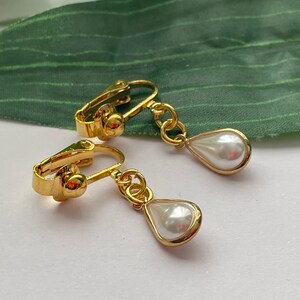 May include: A pair of gold-colored clip-on earrings featuring a teardrop-shaped, white faux pearl pendant. The earrings have a classic design with a polished finish, suitable for various occasions.