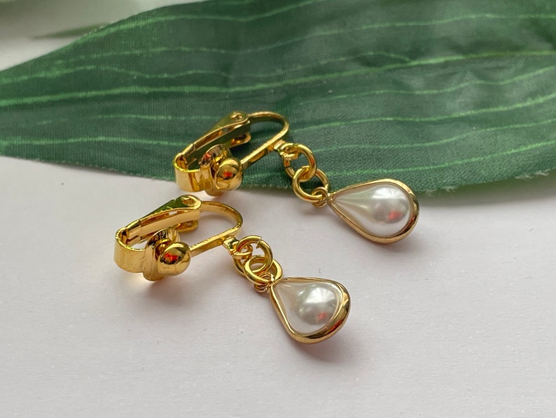 May include: A pair of gold-colored clip-on earrings featuring a teardrop-shaped, white faux pearl pendant. The earrings have a classic design with a polished finish, suitable for various occasions.