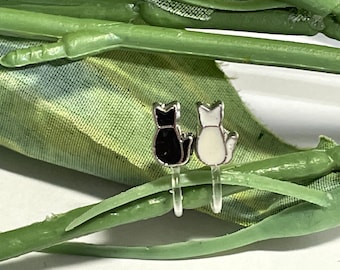 Op de afbeelding: Een paar clip-on oorbellen met kattenontwerpen. Eén oorbel is zwart, de andere wit, beide met zilverkleurige contouren. De oorbellen zijn bevestigd aan doorzichtige plastic clips, tegen een achtergrond van groene en bedrukte stof.