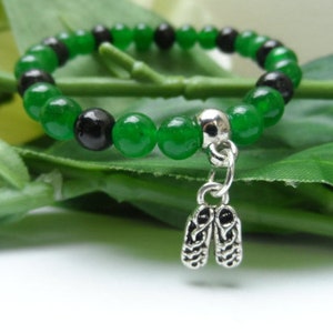 May include: A green and black beaded bracelet with a silver charm in the shape of lungs. The bracelet is on a green leafy background.