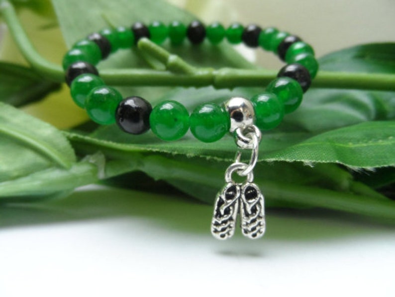 May include: A green and black beaded bracelet with a silver charm in the shape of lungs. The bracelet is on a green leafy background.