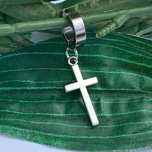 May include: A silver cross charm hangs from a silver ear cuff. The ear cuff is made of stainless steel and has a black finish. The cross charm is a simple design with a rounded top and bottom.