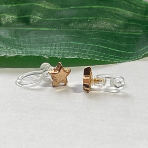 Op de afbeelding: Een paar gouden oorclips in de vorm van een ster met doorzichtige plastic sluitingen.