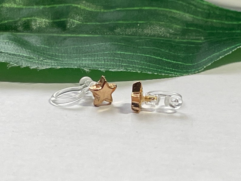 Op de afbeelding: Een paar gouden oorclips in de vorm van een ster met doorzichtige plastic sluitingen.