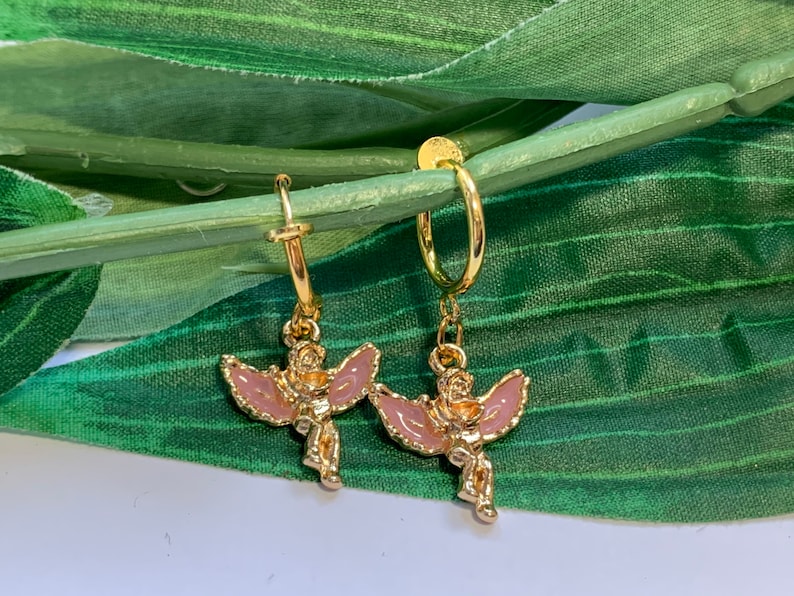 May include: A pair of gold hoop earrings with a pink angel charm dangling from each hoop.