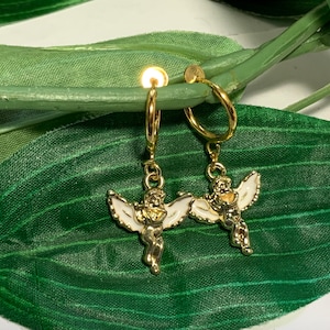 May include: A pair of gold hoop earrings with a white angel charm hanging from each hoop. The angel charms have gold wings and are holding a heart.