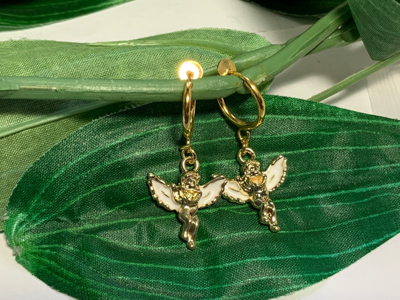 May include: A pair of gold hoop earrings with a white angel charm hanging from each hoop. The angel charms have gold wings and are holding a heart.