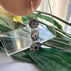 May include: Silver dangle earrings with three round charms. Each charm is decorated with a black, white, and multi-colored enamel design.