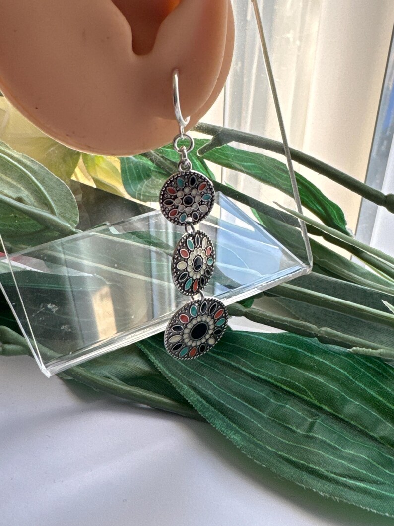 May include: Silver dangle earrings with three round charms. Each charm is decorated with a black, white, and multi-colored enamel design.