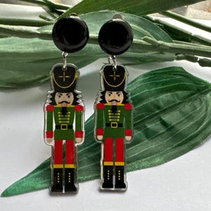 May include: A pair of dangle earrings featuring a nutcracker soldier design. The earrings are made of clear acrylic and have a black round top. The nutcracker soldiers are wearing green uniforms with red accents and black hats.