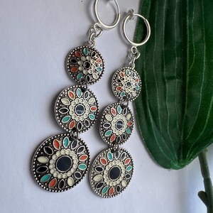 May include: A pair of silver clip-on earrings with three round, colorful, enamel-painted charms. The charms feature a black center with a white and multi-colored floral design.