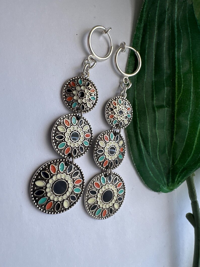 May include: A pair of silver clip-on earrings with three round, colorful, enamel-painted charms. The charms feature a black center with a white and multi-colored floral design.