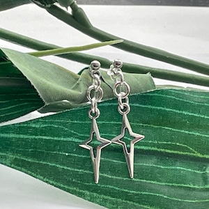 May include: A pair of silver star earrings with a delicate chain. The earrings are hanging from a green leaf background.