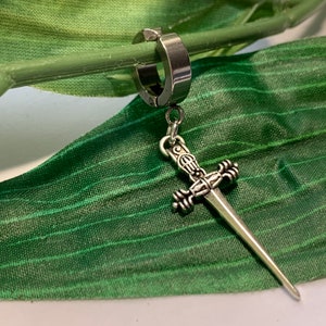 May include: A silver sword charm hanging from a silver hoop earring. The earring is on a green leaf background.