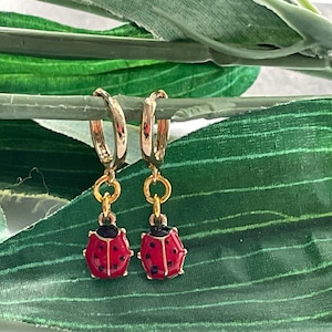 May include: A pair of gold hoop earrings with a dangling ladybug charm. The ladybugs are red with black spots and gold accents.