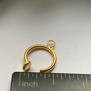 Puede incluir: Un hallazgo de joyer&iacute;a en forma de c&iacute;rculo abierto, de color dorado, con un peque&ntilde;o lazo en la parte superior y un extremo plano y circular. El di&aacute;metro es de aproximadamente 1,3 cm. Una regla est&aacute; en primer plano.