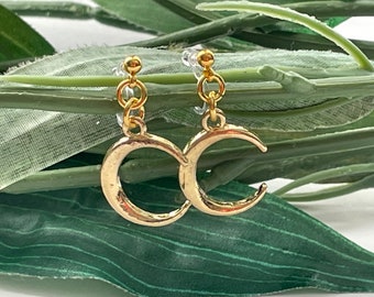 Op de afbeelding: Een paar gouden halve maan oorbellen met een delicate ketting die aan elke oorbel hangt. De oorbellen bevinden zich op een groene, bladerige achtergrond.