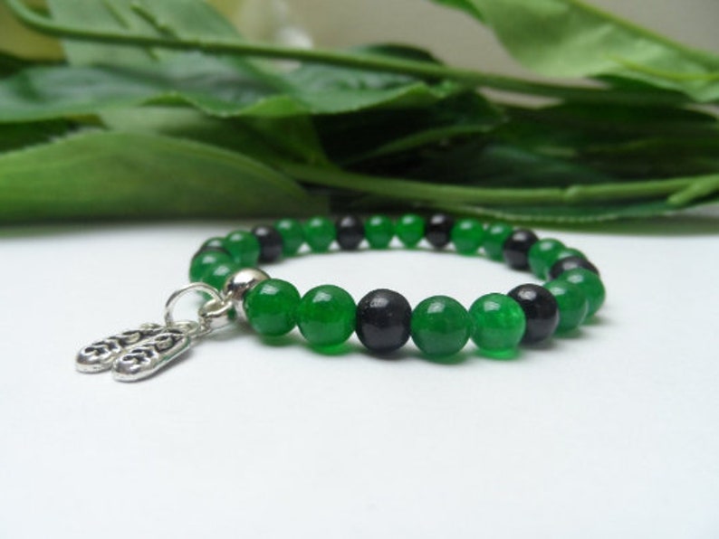 May include: A green and black beaded bracelet with a silver charm. The charm is shaped like two small shoes.