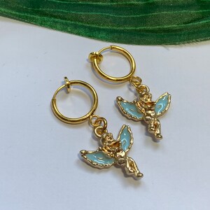 May include: A pair of gold-tone clip-on earrings featuring a small angel charm with light blue wings. The angel is holding a small heart in its hands.