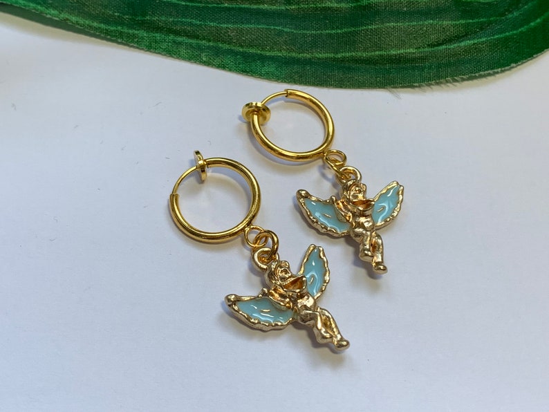 May include: A pair of gold-tone clip-on earrings featuring a small angel charm with light blue wings. The angel is holding a small heart in its hands.