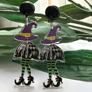 May include: A pair of dangle earrings featuring a witch with a black and white striped skirt, green and black striped stockings, and a purple hat with a green brim. The earrings have black round tops.