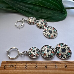 May include: A pair of silver earrings with three round, colorful, patterned charms. The charms are connected by a silver chain. The earrings are measured on a ruler in centimeters.