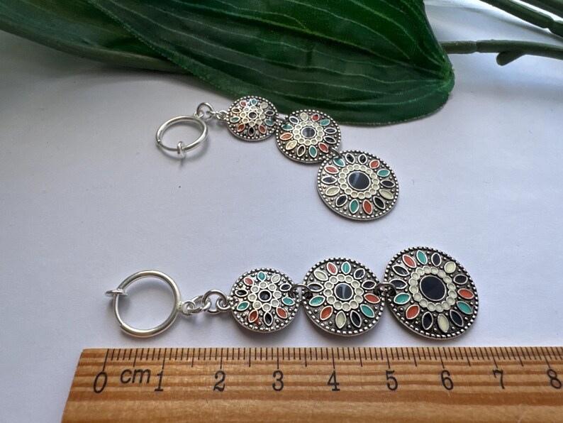 May include: A pair of silver earrings with three round, colorful, patterned charms. The charms are connected by a silver chain. The earrings are measured on a ruler in centimeters.