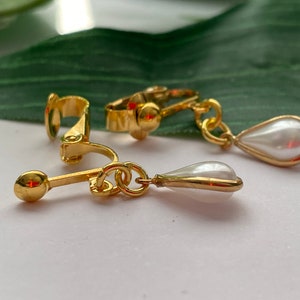 May include: Gold-toned clip-on earrings with a pear-shaped, white faux pearl drop. The earrings have a classic design with a polished finish. The clip-on mechanism is visible, and the earrings are set against a white surface.
