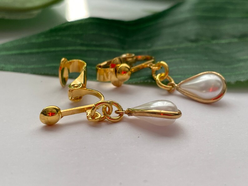 May include: Gold-toned clip-on earrings with a pear-shaped, white faux pearl drop. The earrings have a classic design with a polished finish. The clip-on mechanism is visible, and the earrings are set against a white surface.