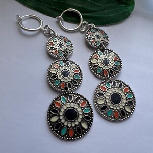 May include: A pair of silver hoop earrings with three dangling charms. Each charm is a round disc with a black center and a white, orange, and green floral design.
