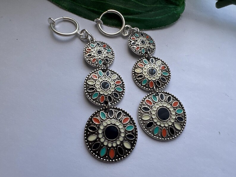 May include: A pair of silver hoop earrings with three dangling charms. Each charm is a round disc with a black center and a white, orange, and green floral design.