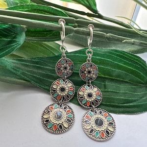 May include: A pair of silver dangle earrings with three round, colorful, patterned charms. The charms have a black center and are decorated with white, green, orange, and blue enamel.