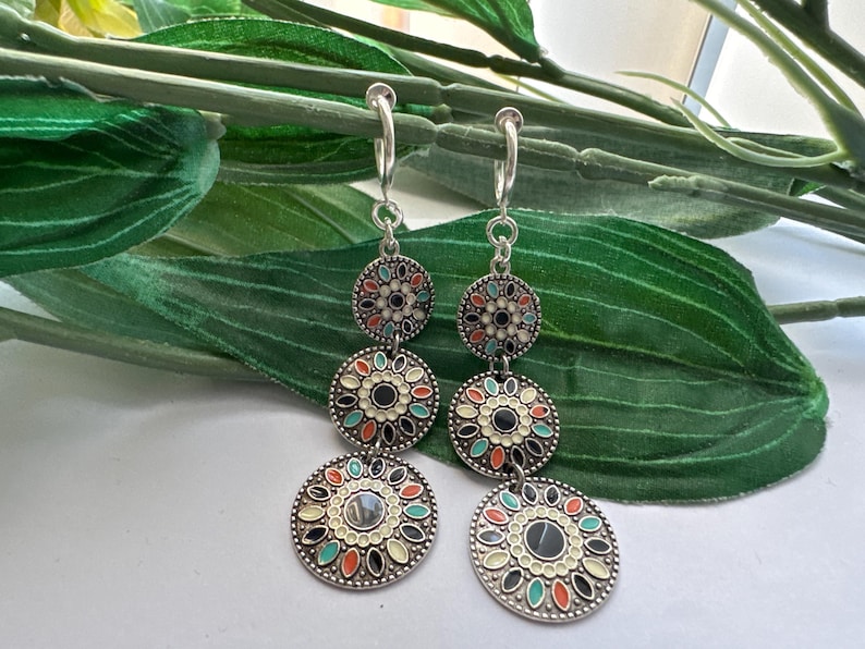 May include: A pair of silver dangle earrings with three round, colorful, patterned charms. The charms have a black center and are decorated with white, green, orange, and blue enamel.